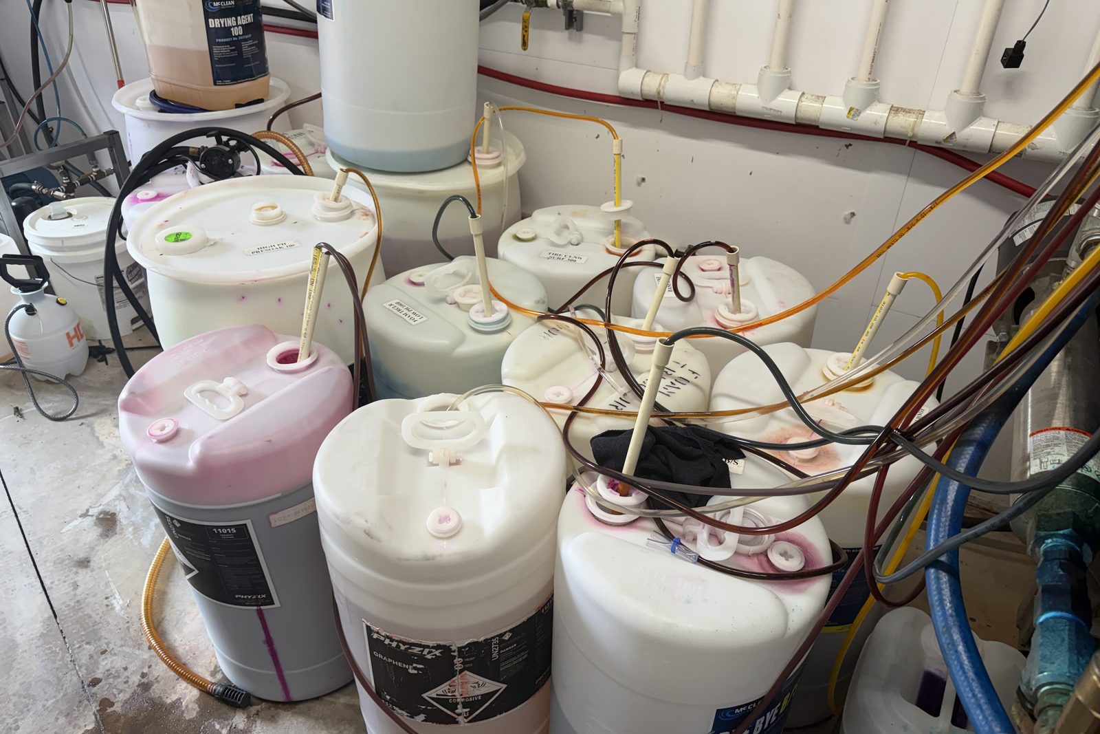Car wash chemical room before the install, with drums and jugs crowded together and dip tubes and hoses running across the floor